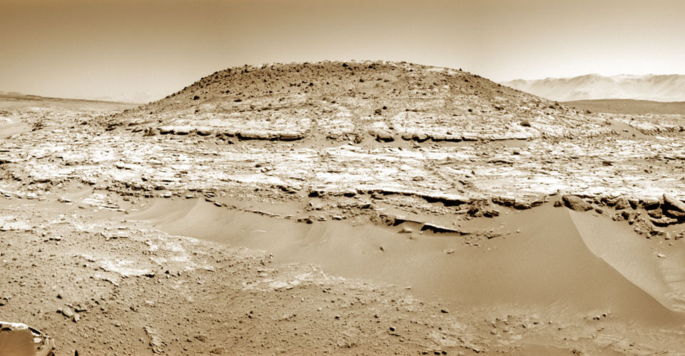 Hinter einer Sandwehe ragt ein 5 Meter hoher Hügel auf. Es ist Mount Remarkable, der Rover Curiosity fand ihn auf dem Weg zu Mount Sharp. Das sepiafarbene Bild zeigt, dass der Hügel waagrecht geschichtet ist.