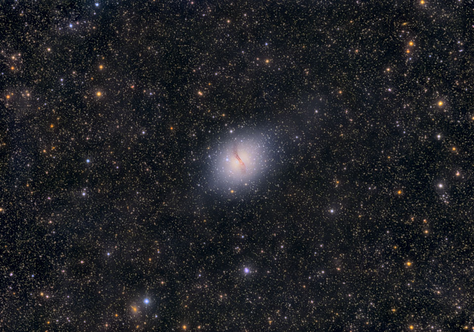 Mitten im Bild leuchtet ein verschwommener heller Fleck mit einem markanten Staubstrang, der über das Zentrum verläuft. Im Hintergrund sind unterschiedlich helle Sterne verteilt.