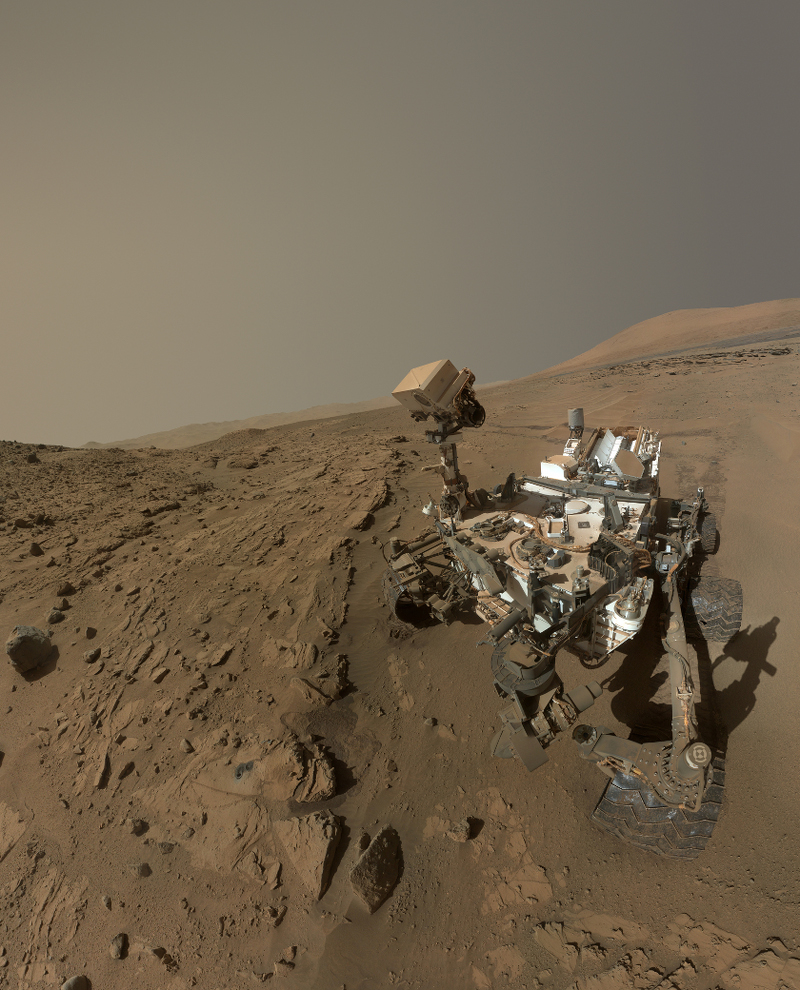 Der Rover Curiosity hockt am Rand eines Geröllfeldes. Er ist nach dem ersten Jahr auf dem Mars von rotem Staub bedeckt. Der Roboterarm, mit dem das Bild fotografiert wurde, ist nicht im Bild.