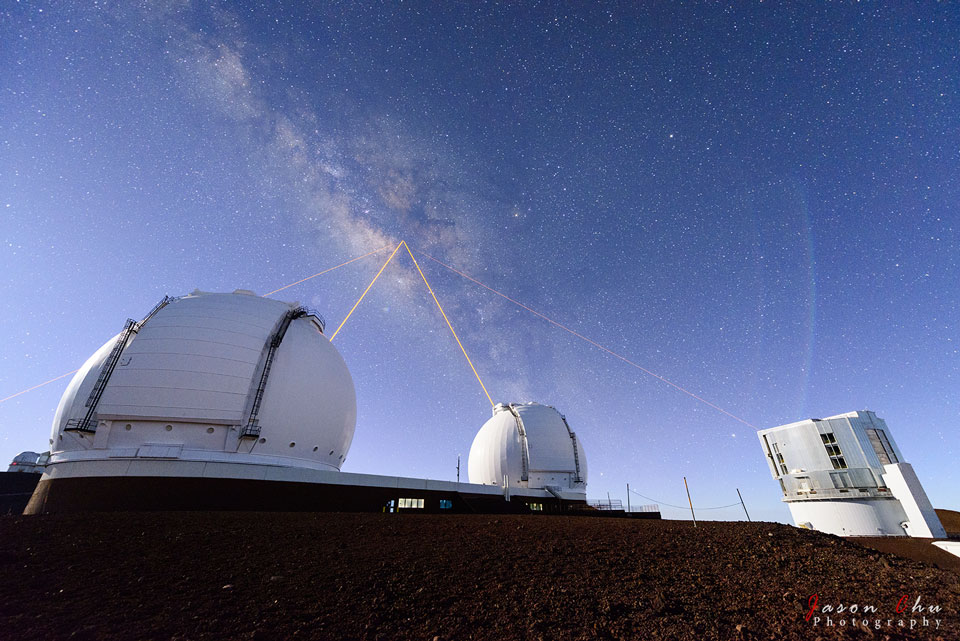 Drei Kuppeln von Sternwarten schicken Laserstrahlen zum Zentrum der Galaxis am Himmel, ein vierter Laserstrahl leuchtet von außerhalb des Bildes hinauf.