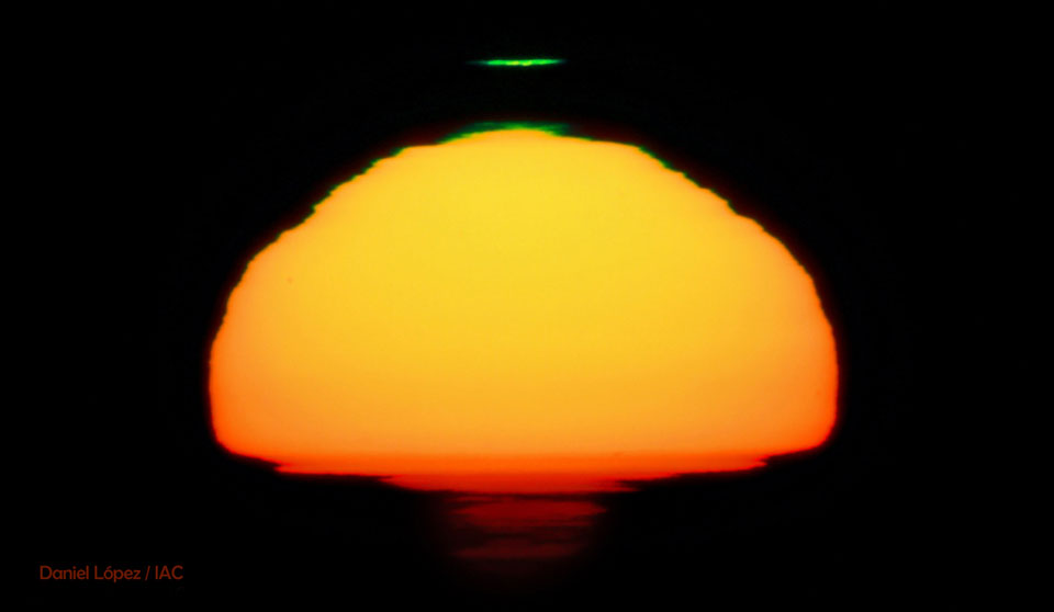 Die untergehende Sonne im Bild ist stark verzerrt durch die Lufthülle der Erde, über der dunkelgelben Form mit rötlichem Rand befindet sich ein schmaler, grün leuchtender Strich.