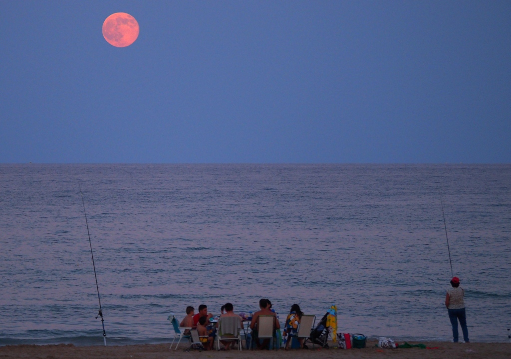 Eine Familie picknickt am Strand von Alicante in Spanien. Zwei Angeln hängen ins Meer, oben leuchtet der rötliche Vollmond im Perigäum größer als sonst.