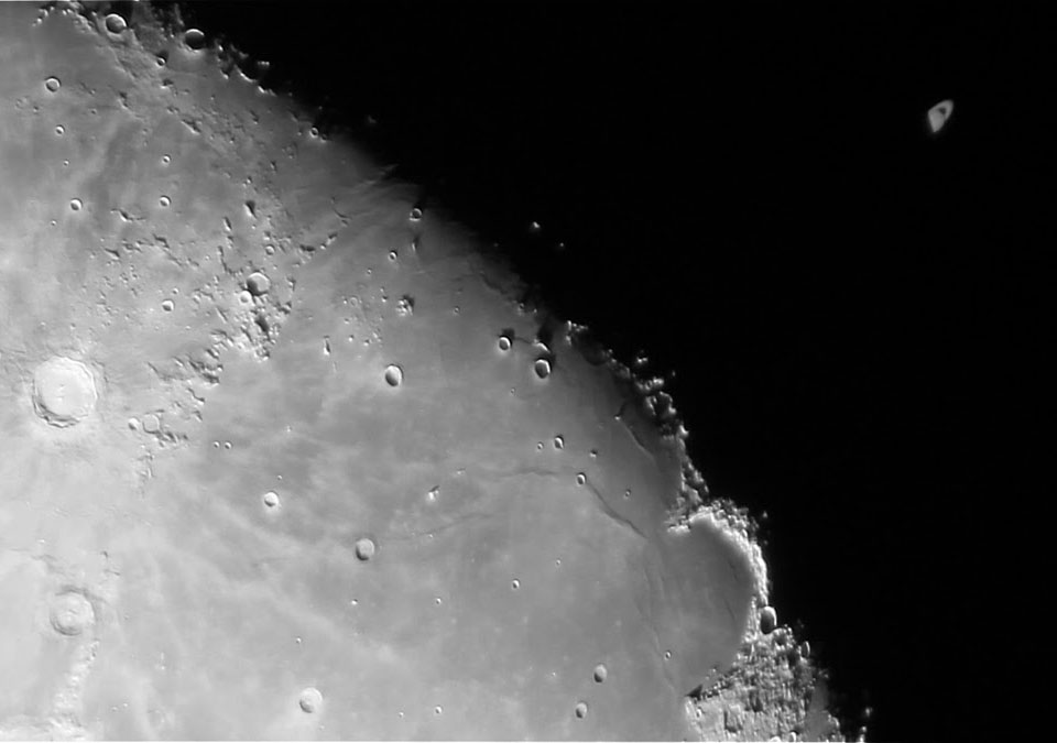 Links unten ist ein Teil des Mondes zu sehen. Diagonal durchs Bild verläuft der Terminator. Rechts oben verschwindet Saturn gerade zur Hälfte hinter dem dunklen Mondrand.