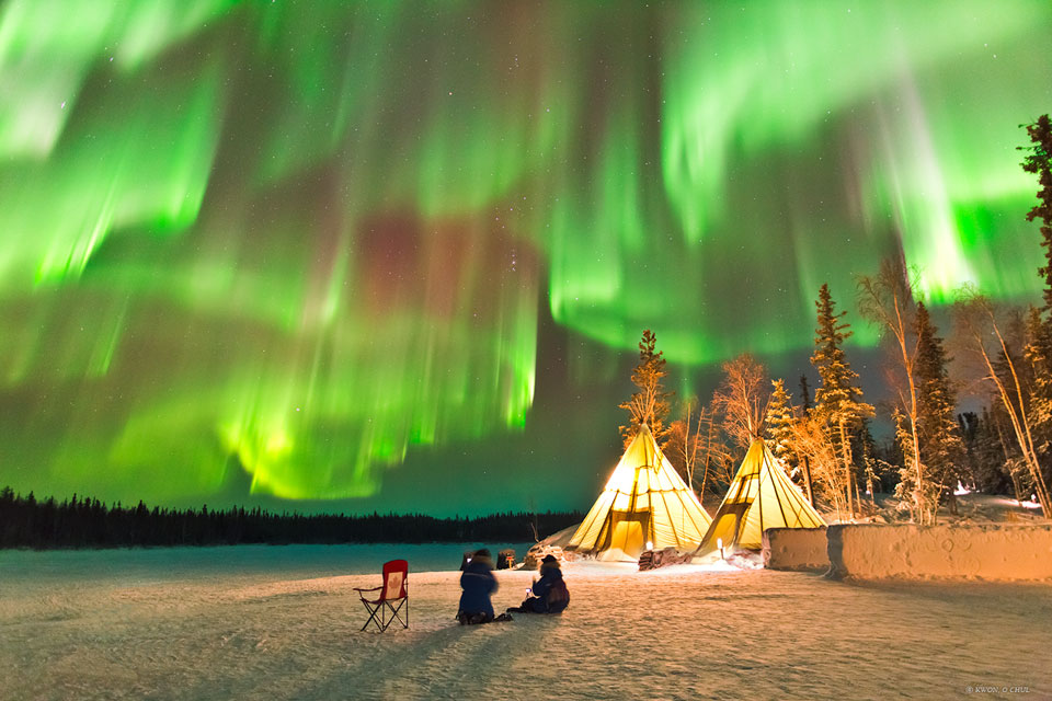 Über zwei Tipis im Schnee wabern Schleier von Polarlichtern. Vor den Tipis sitzen zwei Personen, hinten am Horizont steht ein Wald. Mitten am Himmel leuchtet das Sternbild Orion.