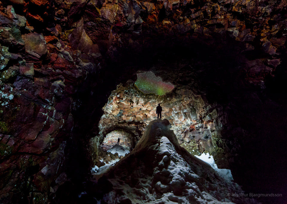 Die isländische Lavahöhle Raufarhólshellir ist innen beleuchtet. Oben ist eine Luke, durch die ein rotes und grünes Polarlicht schimmert.