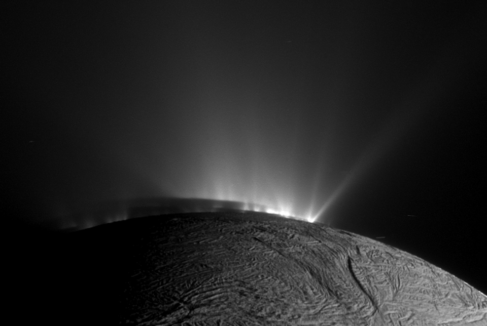 Ein kleiner Teil des Saturnmondes Enceladus ragt vorne auf. Am oberen Rand des Mondes strömt Wasser und Eis ins All. Die Strahlen werden von der Sonne beleuchtet.