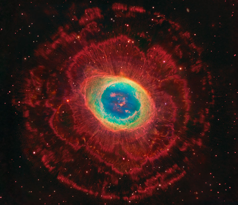 Der Nebel in der Mitte ist der berühmte Ringnebel M57 im Sternbild Leier. Im Bild ist er von riesigen roten Nebellappen umgeben, die mit ihren Strahlen und Fasern an Blütenblätter erinnern.