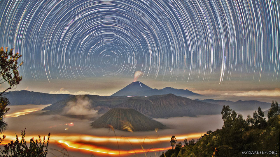 Zwischen den Gipfeln von Vulkanen auf der indonesischen Insel Java leuchten unter dem Nebel die Lichter von Autos. Über den Vulkanen ziehen Sterne ihre kreisförmigen Bahnen um den Himmelspol.