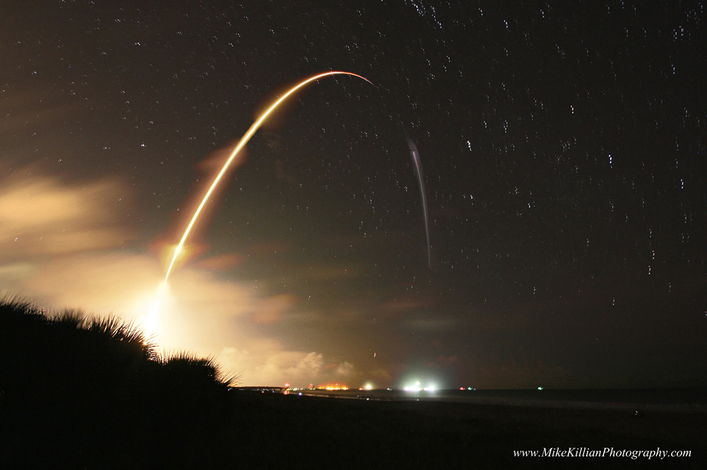 Eine startende Rakete zieht eine glühende Leuchtspur. Der Bogen beginnt links unten und zieht auf einer Parabolbahn über den Himmel, bis er am Horizont verschwidet. Sterne ziehen kurze Strichspuren über Cape Canaveral.