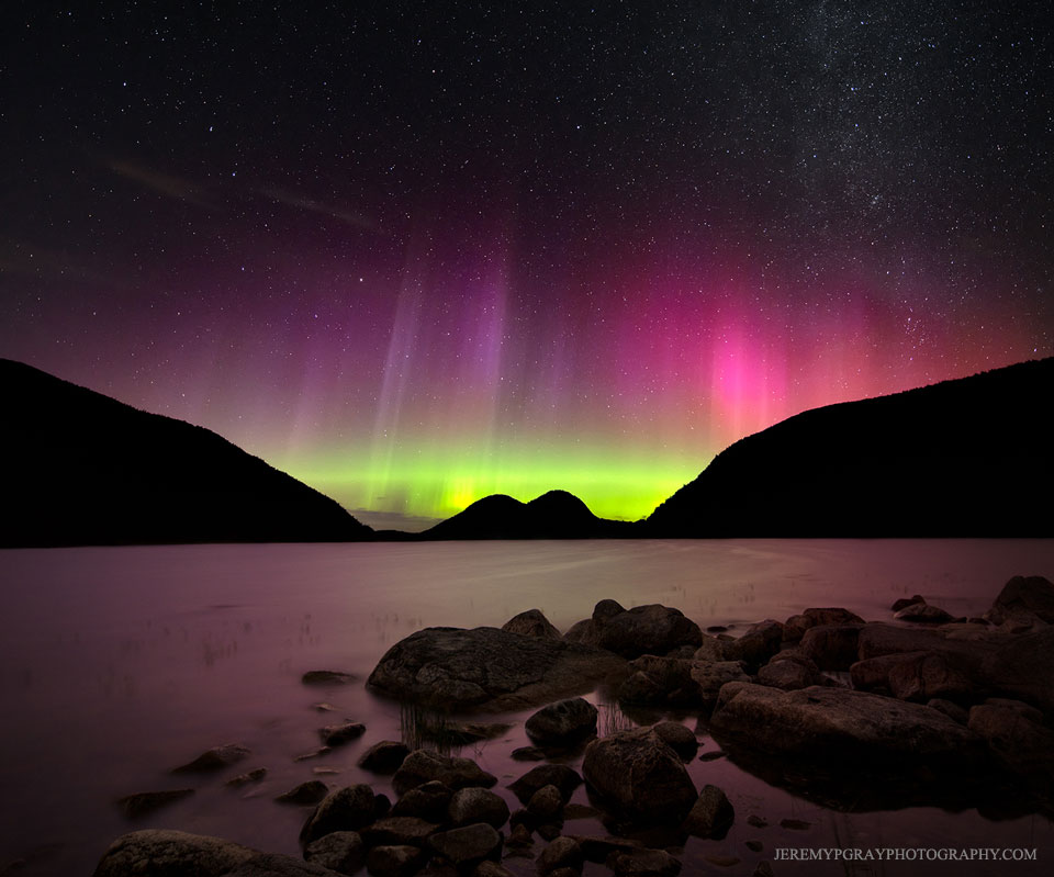 Im Arcadia-Nationalpark in Maine ragen dunkle Silhouetten von Hügeln hinter einem See auf. Am Himmel darüber leuchten grüne und magentafarbene Polarlichter.