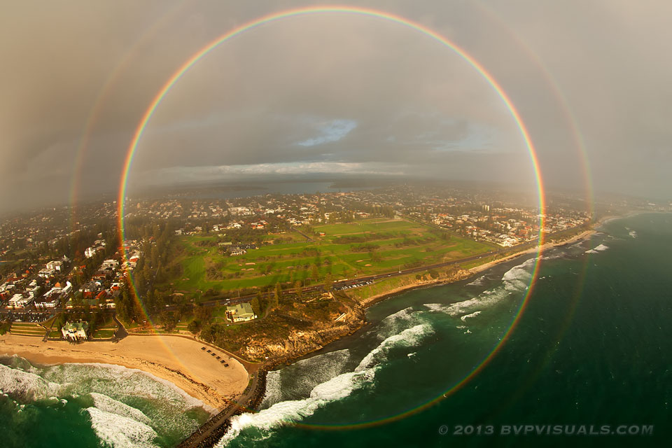 Vor einem Strand mit einer Stadt im Hintergrund leuchtet ein vollständiger 360-Grad-Regenbogen mit einem zweiten Regenbogen außen herum.