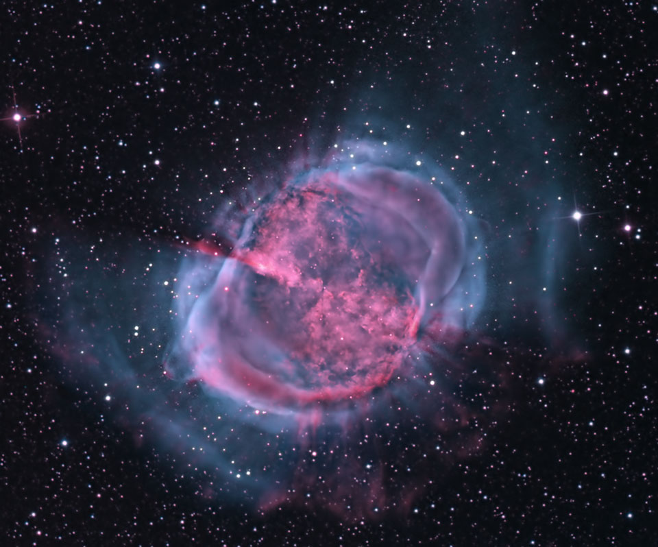 Im Bild leuchtet ein innen zyklamefarbener, außen lila gefärbter Nebel inmitten von Sternen. Seine Form erinnert an eine Hantel. 