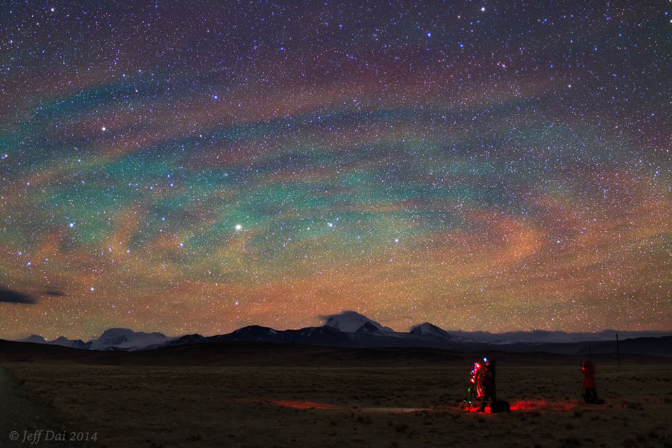 Am Himmel, der von Sternen dicht übersät ist, schimmert buntes Nachthimmellicht in rötlichen und türkisfarbenen konzentrischen Kreisen. Am Boden steht ein rot beleuchteter Astrofotograf mit Teleskop.