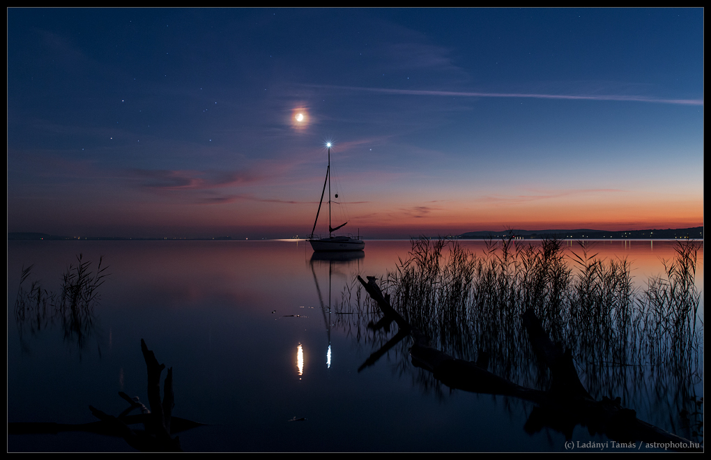 Am ungarischen Balaton spiegeln sich die Lichter von Mond, Antares, Mars, Saturn und dem Positionslicht eines Segelbootes im Wasser. Am Horizont ist noch ein schwaches Abendrot.
