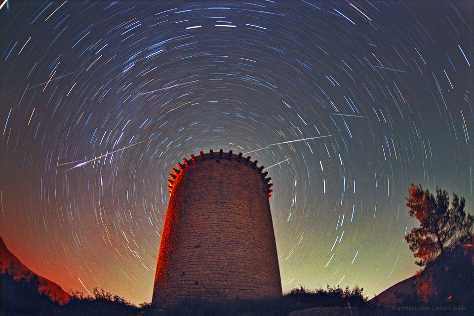 Ein historischer Turm mit Zinnen ist von Strichspuren umgeben, am sternklaren Himmel blitzen Meteore auf. Der Torre de la Guaita ist ein Beobachtungsturm im spanischen Girona. Er stammt aus dem 12. Jahrhundert.