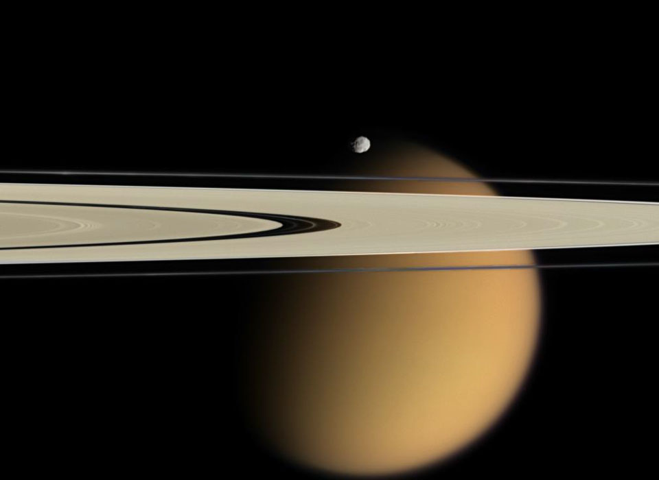 Vor dem wolkigen, ockerfarbenen Mond Titan ragen die Saturnringe von links ins Bild. Sie sind von einer breiten Lücke unterteilt. Über den Ringen schwebt der kleine Mond Epimetheus.