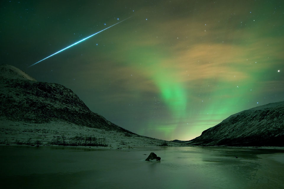 Über der norwegischen Insel Kvaløya bei Tromsø schimmern grüne Polarlichter. Sie beleuchten das eisige Wasser vor verschneiten Berghängen. Links oben zischt eine Feuerkugel der Geminiden über den Himmel.