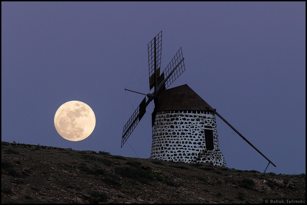 Auf einem Hang steht eine alte Windmühle in der Dämmerung, der Himmel dahinter ist dunkel und graublau. Hinten links geht der Vollmond auf.