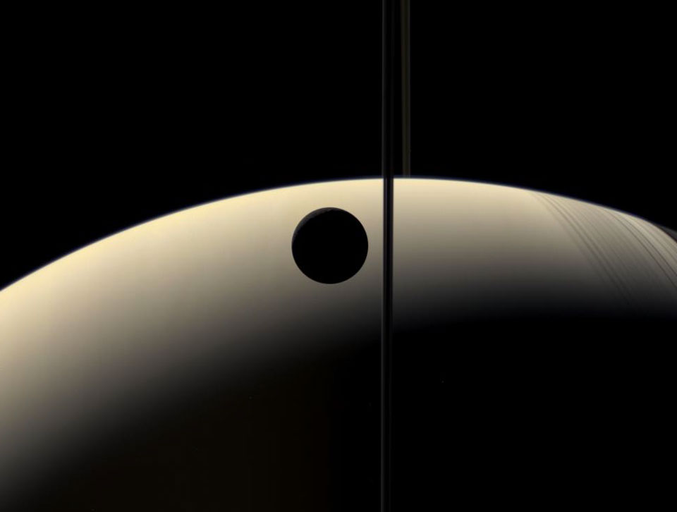 Die beleuchtete Sichel Saturns zeigt nach oben. Durch die Bildmitte verläuft eine senkrechte dunkle Linie, es sind die Saturnringe. Nach rechts werfen sie zarte Schatten. Links neben den Ringen ist der Mond Rhea als dunkler Kreis zu sehen.