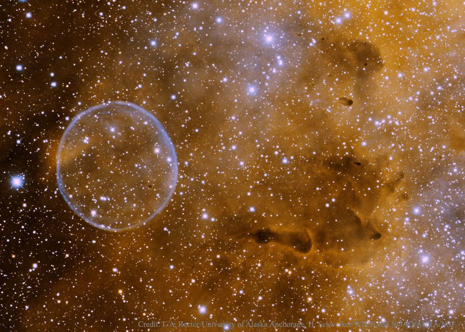 Vor einem Gewimmel aus Sternen und orange-braunen Nebeln zeichnet sich eine Struktur ab, die aussieht wie eine Seifenblase.