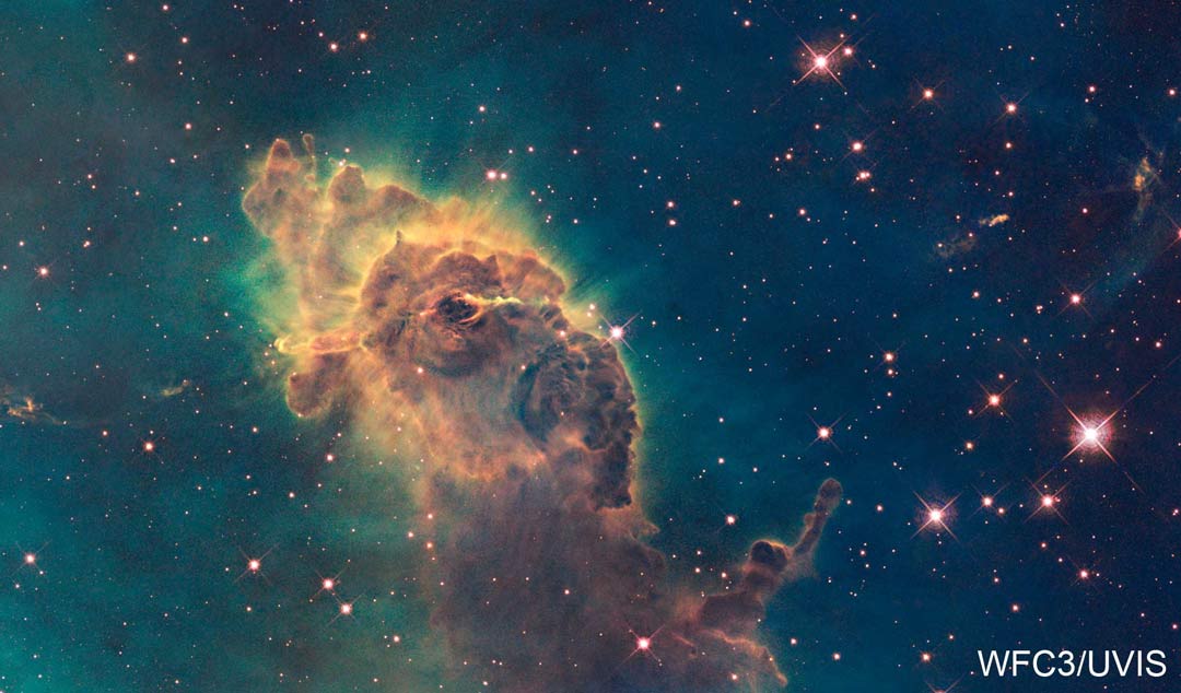 Vor einem petrolfarbenen Hintergrund, der lose mit Sternen übersät ist, ragt eine braune Staubsäule mit sehr starker Struktur auf. Sie ist von hellen gelben Strahlen umgeben.