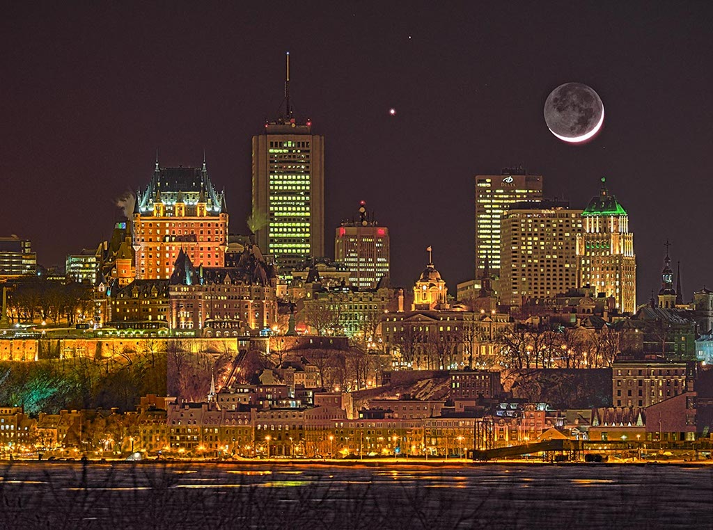 Über der hellen Stadtsilhouette von Quebec bilden der junge Sichelmond und die Planeten Mars und Venus eine hübsche Konjunktion. Auf der dunklen Seite des Mondes sind die Mondmeere erkennbar. Die Stadt leuchtet zu hell, um weitere Sterne zu erkennen.