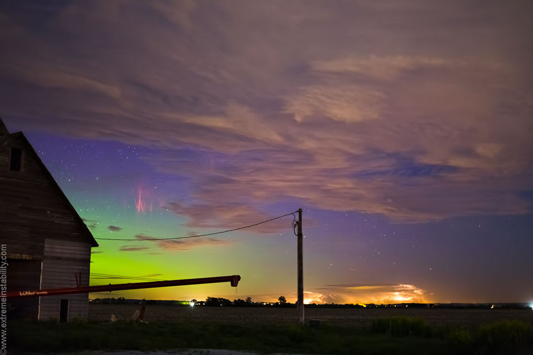 Hinter einem Haus links tobt am Horizont ein Gewitter. Links schimmert ein Polarlicht, darüber zuckt ein prächtiger roter Sprite, auch roter Kobold genannt.