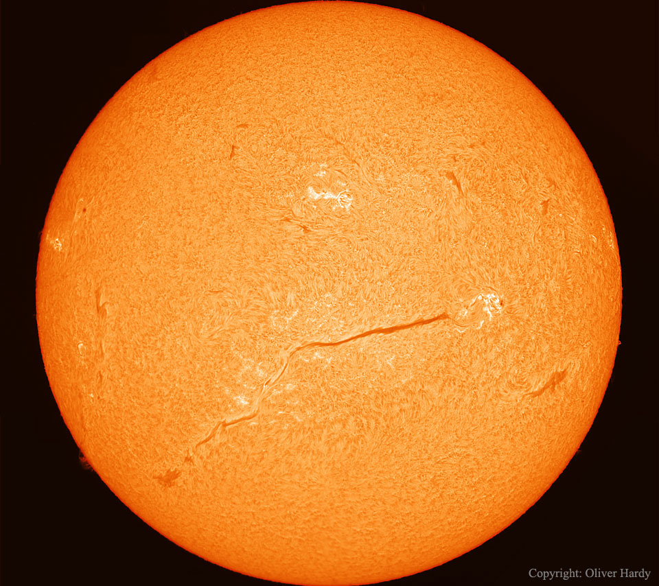 Die Sonne ist bildfüllend dargestellt. Auf der Oberfläche sind pelzartige Strukturen, einige helle Flecken und ein sehr langes dunkles Filament. Am Rand ist die Sonne etwas dunkler und orangefarben.