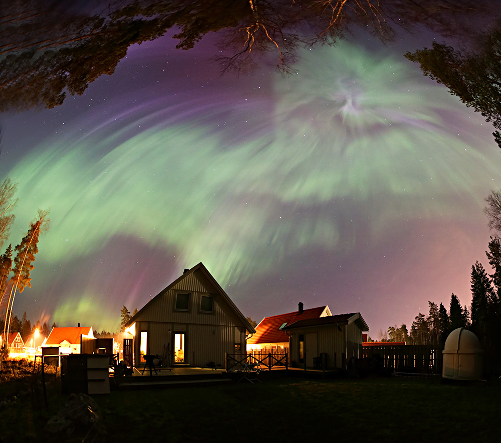 Über einigen Häusern, die nachts beleuchtet sind, wölbt sich der ganze Himmel hoch, der Horizont reicht um das Bild herum und ist bewaldet. Am Himmel leuchtet ein grünes Polarlicht, das vom Zenit ausstrahlt.