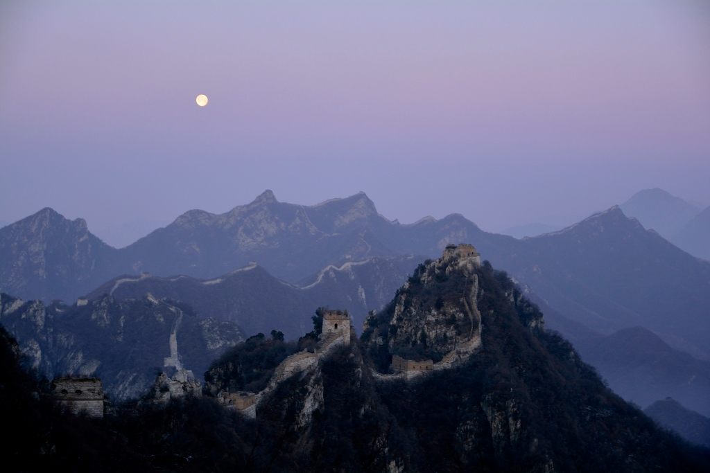 Über den Berggipfeln nördlich von Peking oder Bejing mäandert die Chinesische Mauer durchs Bild. Über den Bergen steigt der Erdschatten auf. An seinem oberen Rand ist der rosarote Venusgürtel. In diesem leuchtet der Mond.