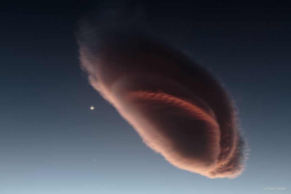 Eine riesige, rosarot beleuchtete Wolke, die an ein Raumschiff erinnert, schwebt am Himmel neben dem winzigen Sichelmond. Kaum erkennbar leuchten weit unter dem Mond die Planeten Venus und Mars.