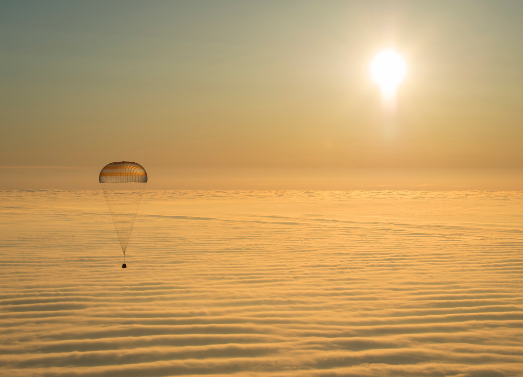 Über einem goldenen Wolkenmeer mit sanften Wellen schwebt ein Fallschirm mit einer Raumkapsel in der Sonne, die gerade aufgegangen ist.
