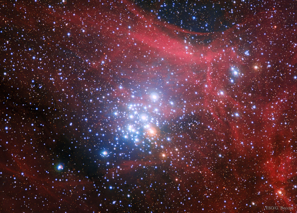 Im Bild ist ein relativ kompakter Sternhaufen, rechts oben breiten sich rote Nebelschleier aus, das ganze Bild ist mäßig von Sternen gefüllt.