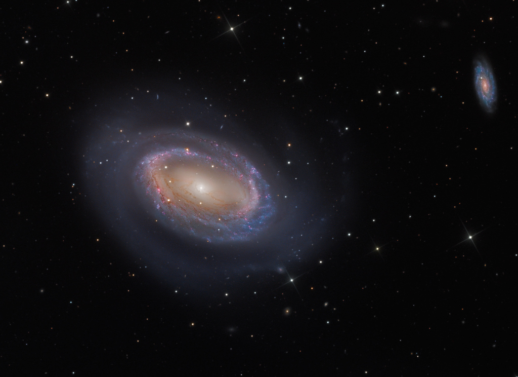 Im Bild schwebt eine Spiralgalaxie, die nur einen einzigen Spiralarm besitzt. Um den gelblichen Kern verläuft ein Ring aus blauen Sternhaufen und rötlichen Sternbildungsregionen. Rechts oben ist eine viel weiter entfernte gewöhnliche Spiralgalaxie. Nur wenige Sterne sind im Bild verteilt.