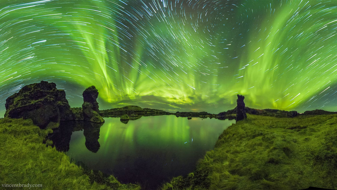 Der spiegelglatte See Mývatn vorne reflektiert Sterne und Polarlichter, die über den Himmel ziehen. Er ist von Lavasäulen umgeben.