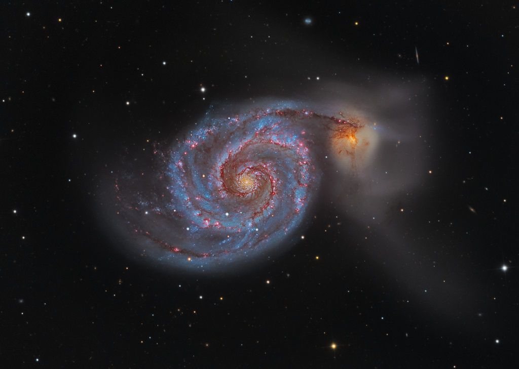 In der Mitte ist eine Spiralgalaxie mit markanten Armen von oben zu sehen, die von vielen blauen Sternen und roten Sternbildungsstrukturen markiert ist. Oben rechts verläuft ein Spiralarm zu einer kleineren Begleitgalaxie, die gelbrötlich leuchtet.