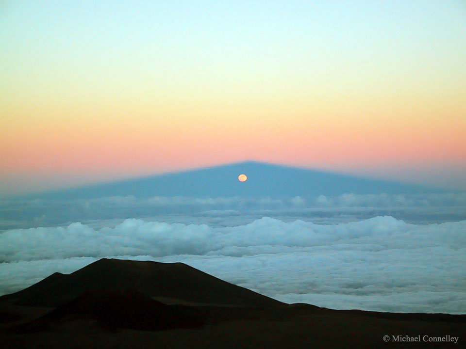 Der Mauna Kea wirft seinen langen Schatten über das Meer. Im Vordergrund ist ein Gipfel zu sehen, hinter den Wolken leuchtet der Mond im Schattenkegel, darüber ist der Erdschatten und ein rötlicher Gürtel zu sehen, der in den blauen Himmel übergeht.