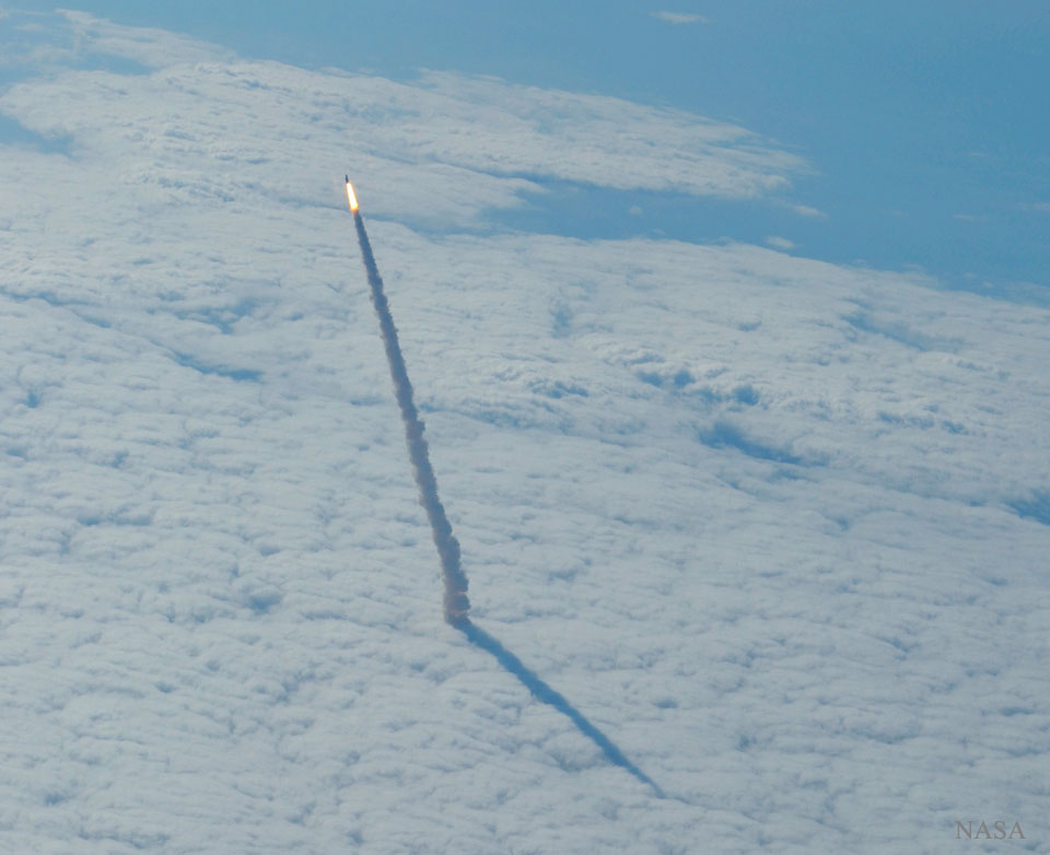 Aus einer Wolkendecke steigt ein Abgasstrahl auf. An seiner Spitze ist der leuchtende Feuerschweif der Raumfähre Endeavour. Der Schatten der Rauchfahne fällt auf die Wolken. Hinten ist ein Ozean zu sehen.