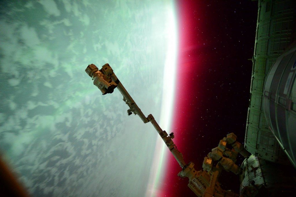 Links schimmert die Erde seltsam grünlich, der Horizont, der in der Mitte senkrecht verläuft, ist rot gesäumt. Rechts ist die Internationale Raumstation. Ein Roboterarm ragt ins Bild.