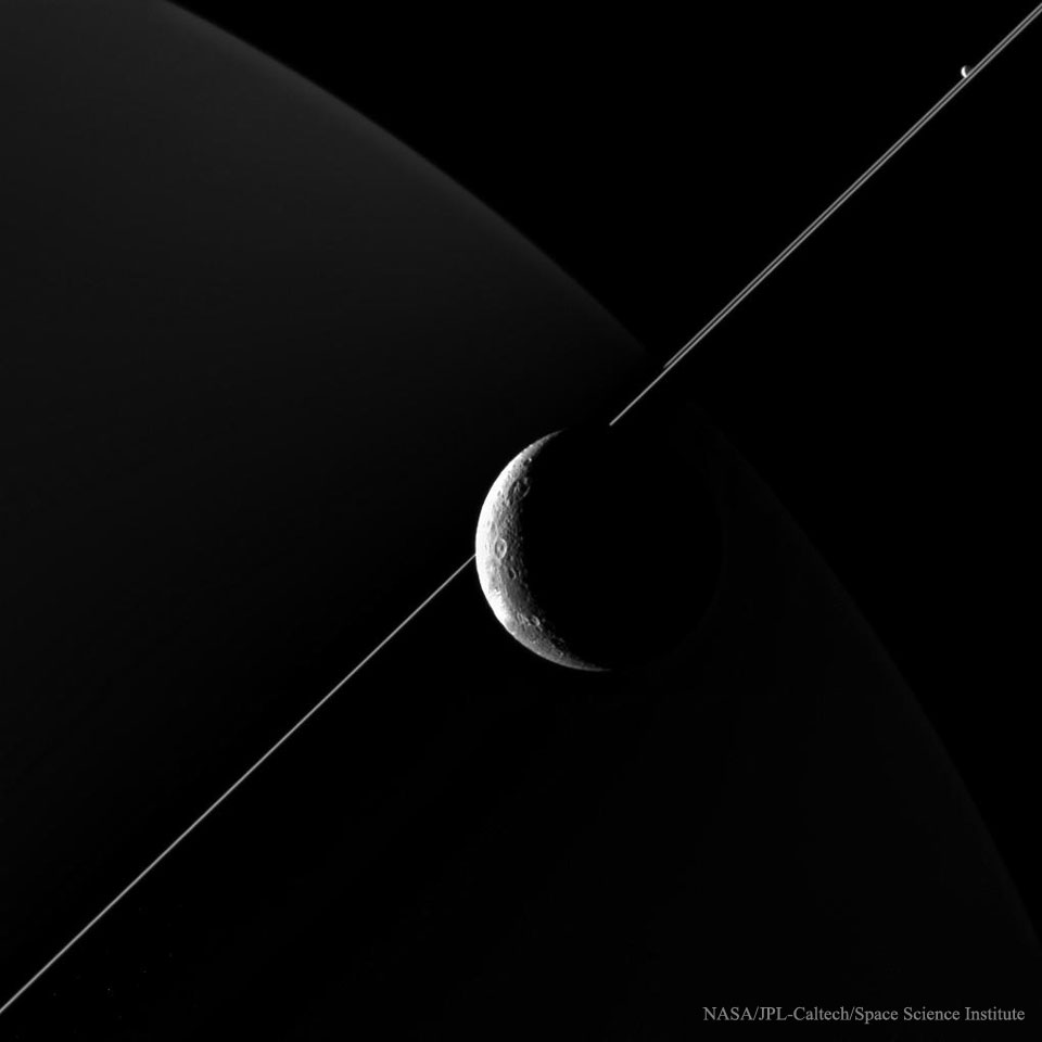 Mitten im Bild leuchtet der Saturnmond Dione. Die Ringe verlaufen diagonal hinter Dione durchs Bild. Links oben ist die schmale Sichel des Planeten Saturn zu sehen, der nicht direkt von der Sonne, sondern indirekt von den Ringen beleuchtet wird.