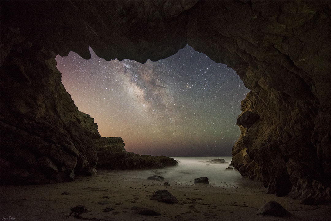 Aus einer Höhle fällt der Blick auf das Heer hinter Sand, der in der Höhle liegt. Über dem Horizont steigt die Milchstraße mit ihren dunklen Staubwolken auf.