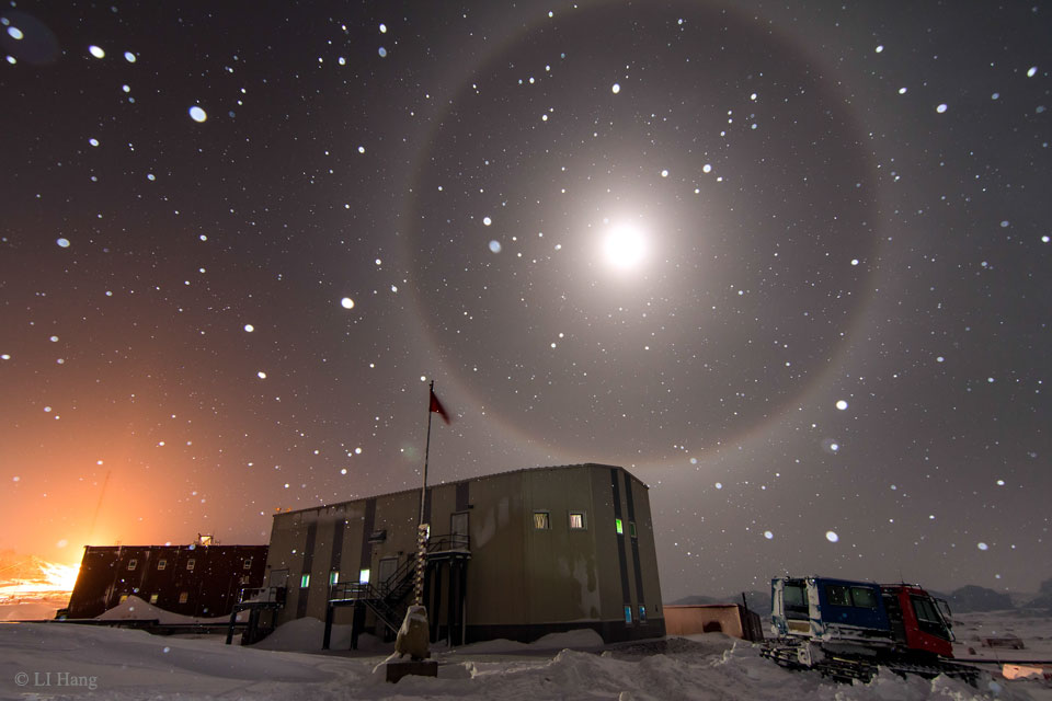 Über der Zhongshan-Station in der Antarktis leuchtet der Mond. Er ist von einem 22-Grad-Halo umgeben. Vorne stehen ein Stromaggregat und ein Schneemobil.