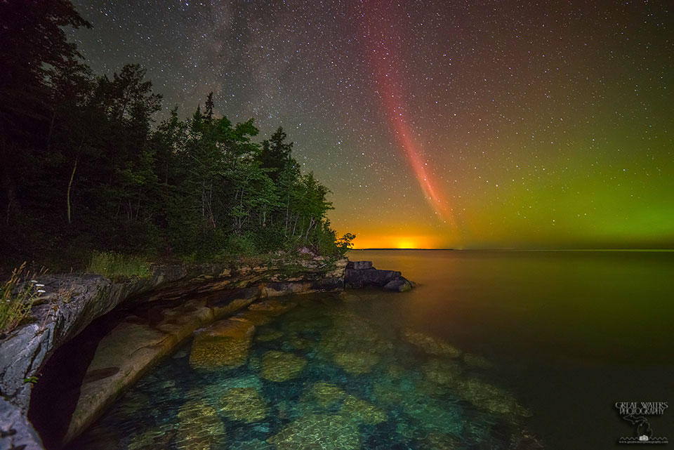 Über dem Lake Superior (Oberer See) leuchtet ein rotes bogenförmiges Licht. Links daneben ist gelbe Lichtverschmutzung am Horizont. Der See liegt rechts, links ist ein Wald an einer felsigen Küste.