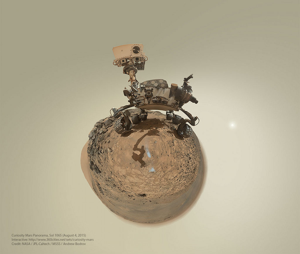 An einem beigefarbigen Himmel mit einer trüben Sonne schwebt ein rostbrauner Planet, auf dem ein Marsrover hockt. Am Boden sieht man den Schatten des Roboterarms, mit dem die Bilder aufgenommen wurden, aus denen das Panorama entstand. Den Arm selbst sieht man nicht.