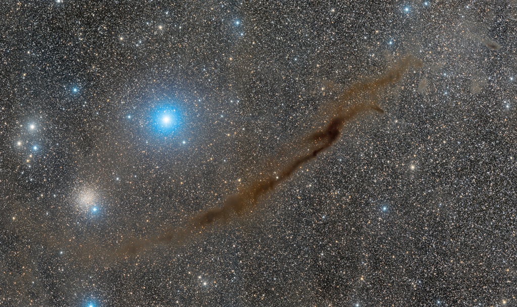 Ein dunkler Nebel verläuft diagonal durchs Bild. Dahinter sind dichte Sternwolken. Links schimmert der weit entfernte Kugelsternhaufen NGC 4372.