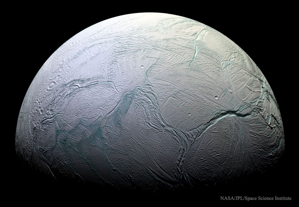 Der Saturnmond Enceladus ist halb beleuchtet. Seine Oberfläche ist von tiefen Schluchten überzogen, die hier teilweise blau gefärbt sind. Die Schattengrenze ist unten.