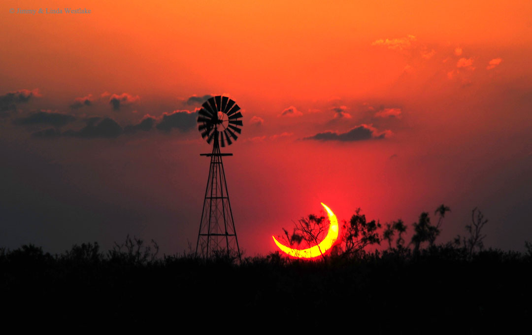 Hinter den Silhouetten von Bäumen, Gestrüpp und einer Windmühle geht eine Sichel unter. Es ist die Sonne, die teilweise vom Mond verdeckt wird. Der Himmel ist leuchtend rot.