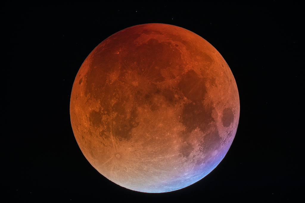 Der Mond ist in den rötlichen Erdschatten getaucht. Rechts unten leuchtet der Rand bläulich, dort fällt Sonnenlicht darauf, das durch die Ozonschicht der Erde gedrungen war.