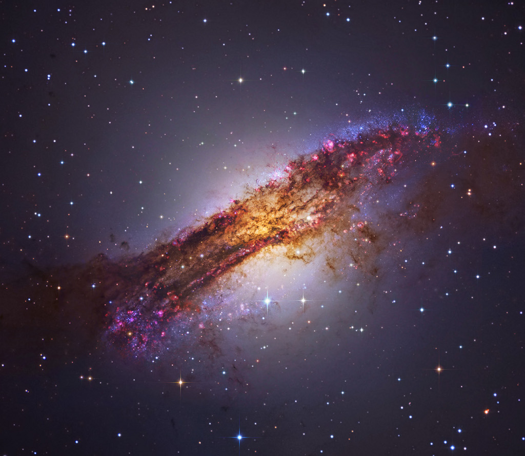 Mitten im Bild leuchtet eine runde verschwommene Struktur, vor der eine sehr markante, breite Staubwolke verläuft, die von rosaroten Sternbildungsregionen und blauen Sternhaufen gesprenkelt ist.