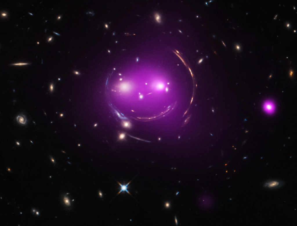 Mitten im Bild grinst ein violettes Gesicht aus Galaxien. Es ist von Bögen eingerahmt. Die Bögen sind die verzerrten Bilder von Galaxien, die weit dahinter liegen.